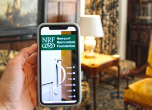 A hand holding a phone with the NRF app homepage on the screen. In the background is the Morning Room at Rough Point, including a 16th century painting, furniture, and patterned curtains